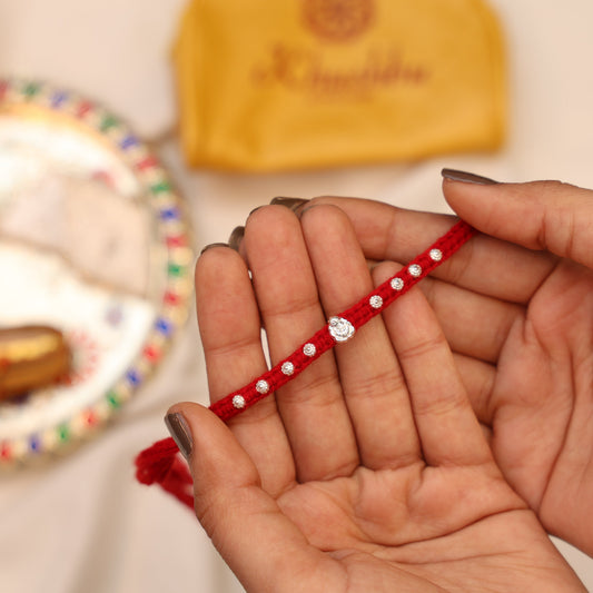 Hand holding a red bracelet with a diamond-like stone, blurred background with decorative items.