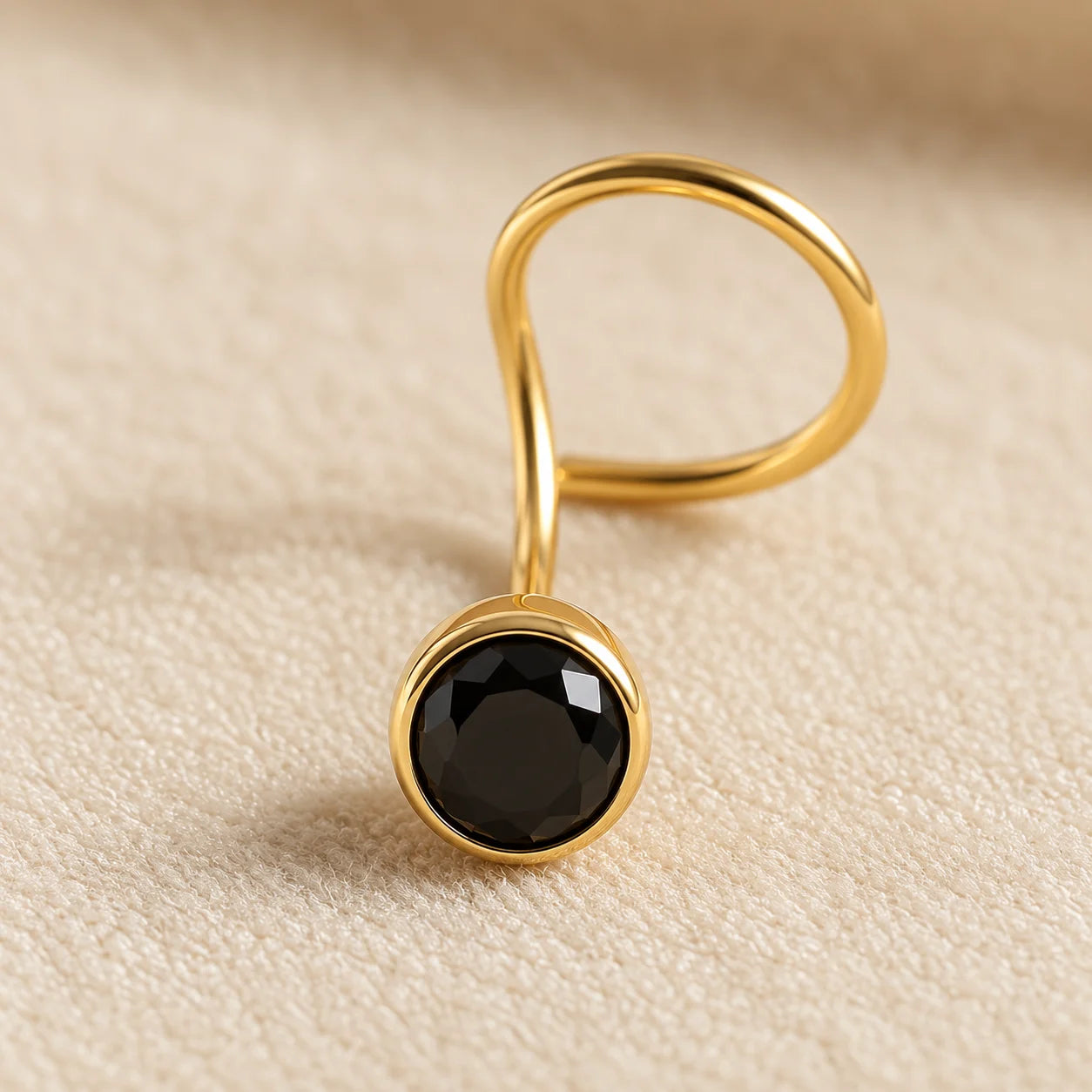 A gold-plated ring with a twisted band and a round bezel-set black faceted gemstone, resting on a textured beige fabric background.