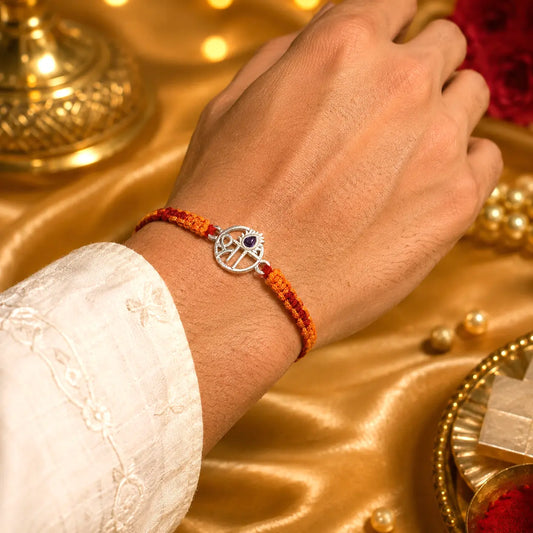 Shree silver rakhi with purple stone and red orange thread on golden satin background