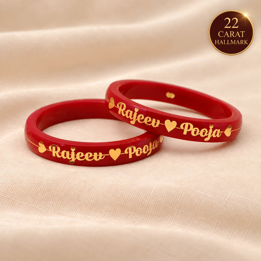 Two red bangles with gold engraved script reading 'Rajeev' and 'Pooja' separated by small hearts, displayed on a beige fabric background. A gold circular badge in the top right says '22 CARAT HALLMARK'.