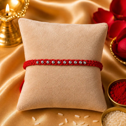 925 Silver Bead Rakhi with handcrafted red thread displayed on a beige velvet cushion with diya, kumkum, rice, and festive Raksha Bandhan decorations.