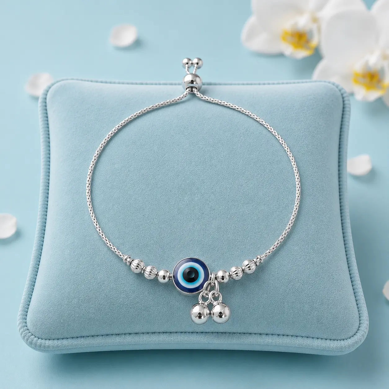 925 Sterling Silver Evil Eye Adjustable Bracelet displayed on a soft blue velvet jewelry cushion with white orchid flowers and luxury studio lighting.