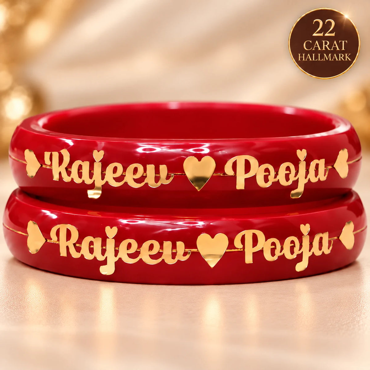 Two red bangles stacked on a light beige surface, each engraved with the names 'Rajeeu' and 'Pooja' separated by a gold heart, with small gold heart accents on the sides. A circular badge in the top right corner reads '22 CARAT HALLMARK'.