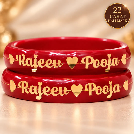 Two red bangles stacked on a light beige surface, each engraved with the names 'Rajeeu' and 'Pooja' separated by a gold heart, with small gold heart accents on the sides. A circular badge in the top right corner reads '22 CARAT HALLMARK'.