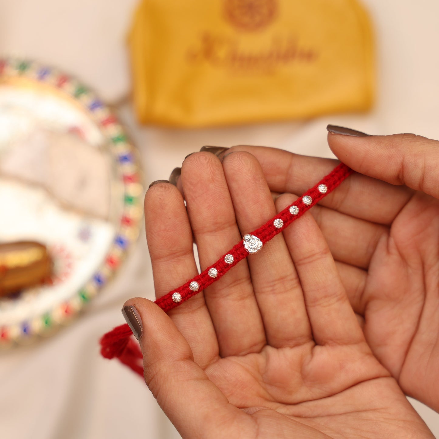 Hand holding a red bracelet with a diamond-like stone, blurred background with decorative items.