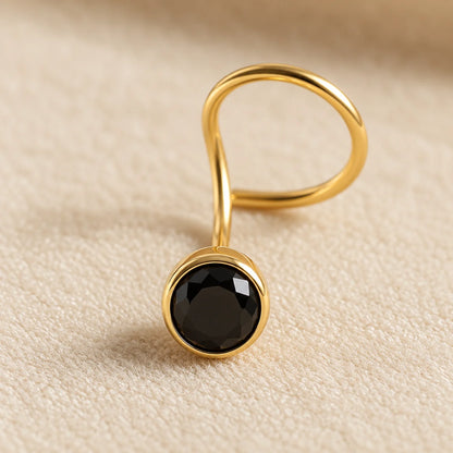 A gold-plated ring with a twisted band and a round bezel-set black faceted gemstone, resting on a textured beige fabric background.