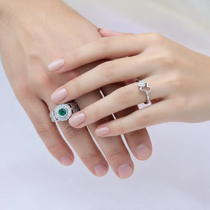 Two hands with pale pink manicured nails wearing silver rings. The left hand wears a silver ring with a round green gemstone surrounded by small clear stones, and the right hand wears a silver ring with a small clear heart-shaped gemstone.