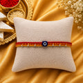 Premium handmade Evil Eye Rakhi with blue protective bead on a cream cushion, luxury golden festive background with kumkum, rice, sweets, and flowers for Raksha Bandhan.