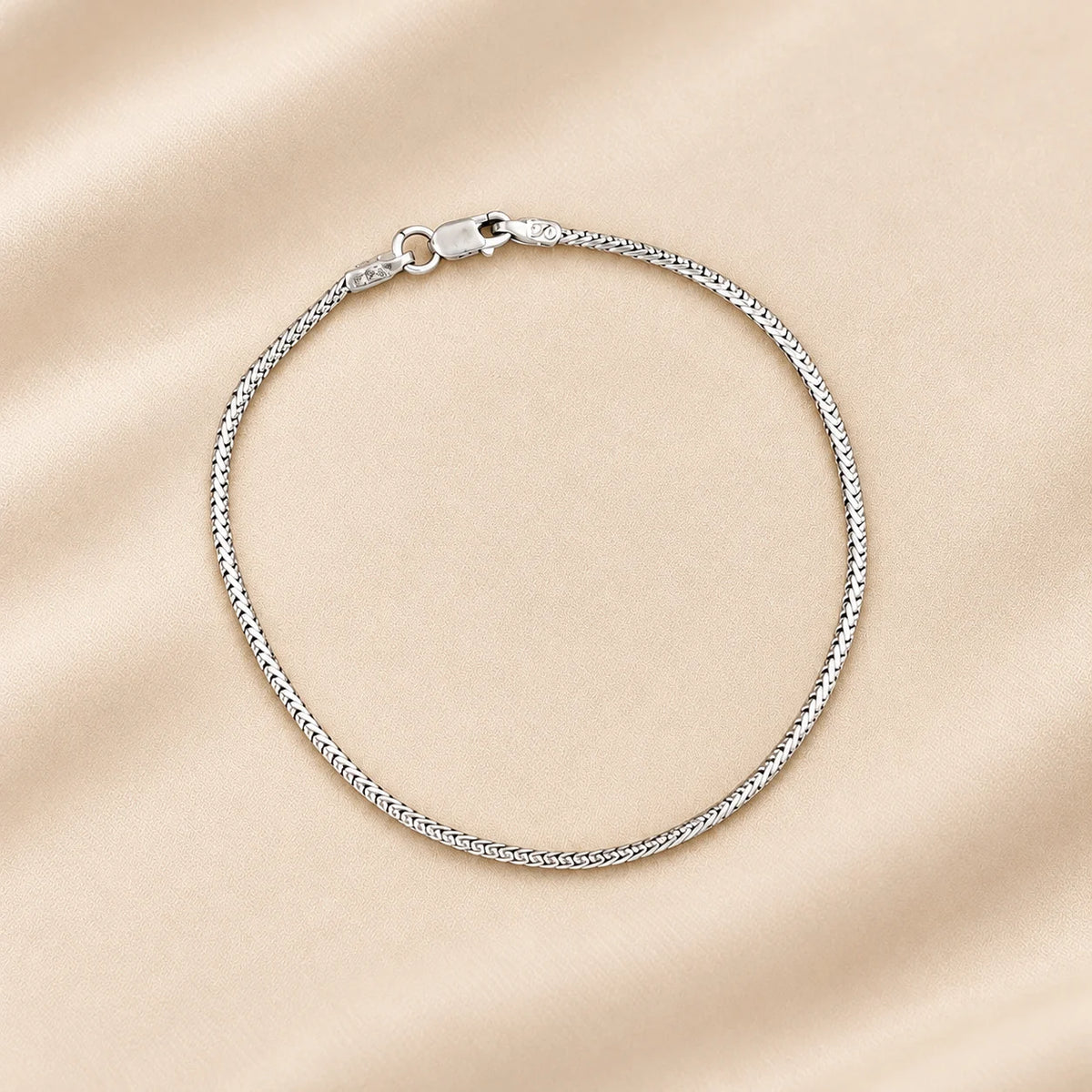 A silver chain bracelet with a textured, rope-like link design and a lobster clasp, displayed on a soft beige satin fabric background.