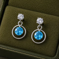 Silver blue round cubic zirconia open circle drop earrings for women with polished silver finish and sparkling crystal accents displayed on a white rose branch.