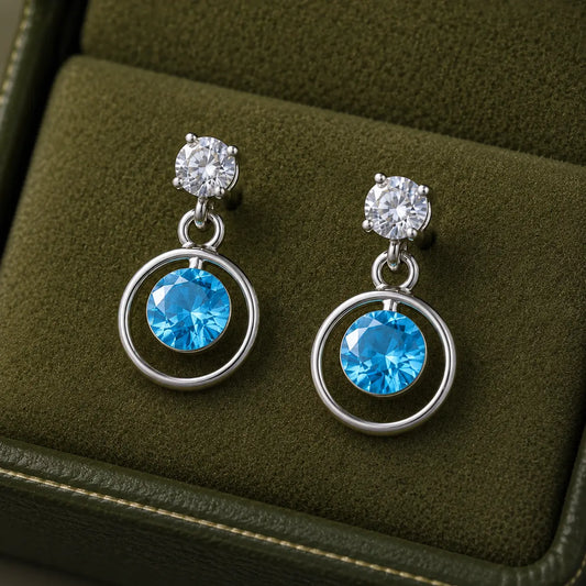Silver blue round cubic zirconia open circle drop earrings for women with polished silver finish and sparkling crystal accents displayed on a white rose branch.