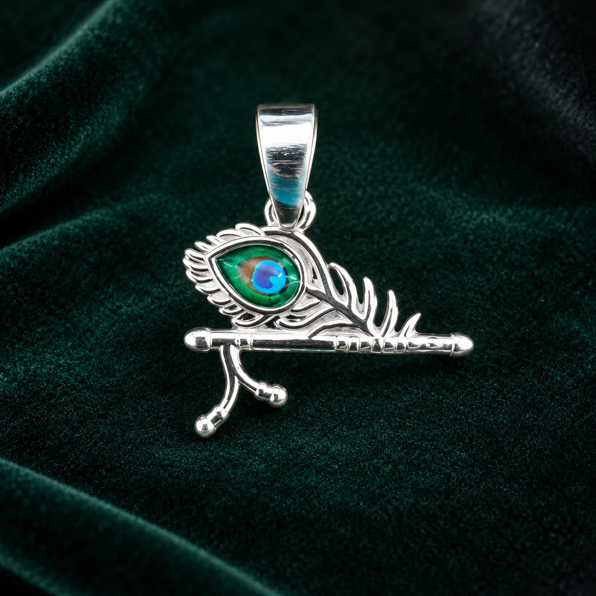 Silver Krishna flute peacock feather pendant with blue and green enamel detailing on a dark teal velvet background