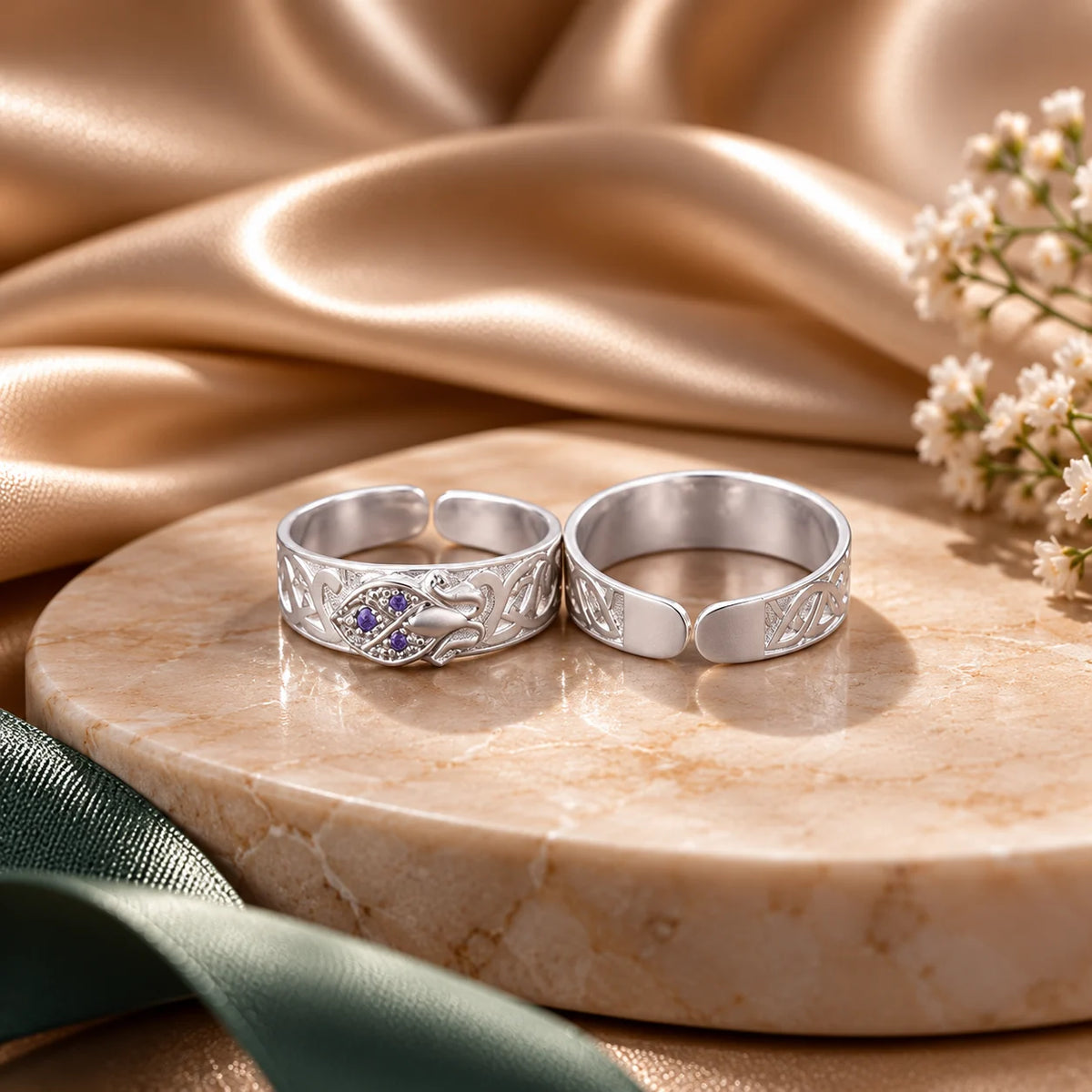 Two silver-toned adjustable rings displayed on a round beige stone slab, with draped champagne satin fabric, a green ribbon, and small white flowers in the background.