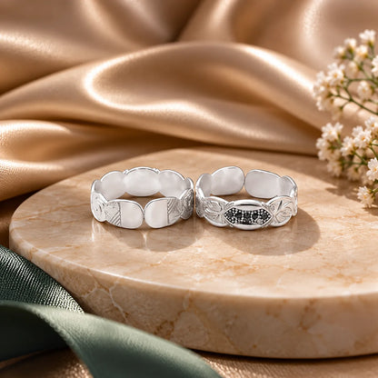 Two silver rings with decorative leaf and petal motifs, one plain and one with a small black-and-white floral center, displayed on a round beige marble tray with draped satin fabric and white flowers in the background.