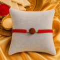 Premium handmade Rudraksha Rakhi with natural Rudraksha bead, silver spacer beads, and red braided thread displayed on a grey velvet cushion with a luxurious Raksha Bandhan festive background featuring kumkum, rice, and golden satin fabric.