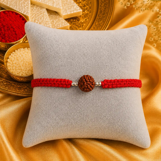 Premium handmade Rudraksha Rakhi with natural Rudraksha bead, silver spacer beads, and red braided thread displayed on a grey velvet cushion with a luxurious Raksha Bandhan festive background featuring kumkum, rice, and golden satin fabric.