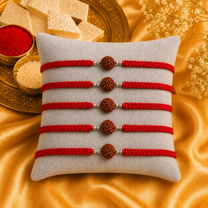 Premium handmade Rudraksha Rakhi with natural Rudraksha bead, silver spacer beads, and red braided thread displayed on a grey velvet cushion with a luxurious Raksha Bandhan festive background featuring kumkum, rice, and golden satin fabric.