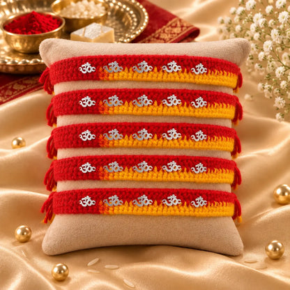 "Set of 5 handcrafted red and yellow thread Rakhis with silver Om charms displayed on a beige velvet cushion, perfect for Raksha Bandhan celebrations."