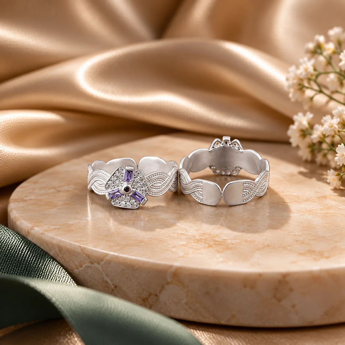 Two silver rings displayed on a round beige marble tray, with draped champagne satin fabric and small white flowers in the background.