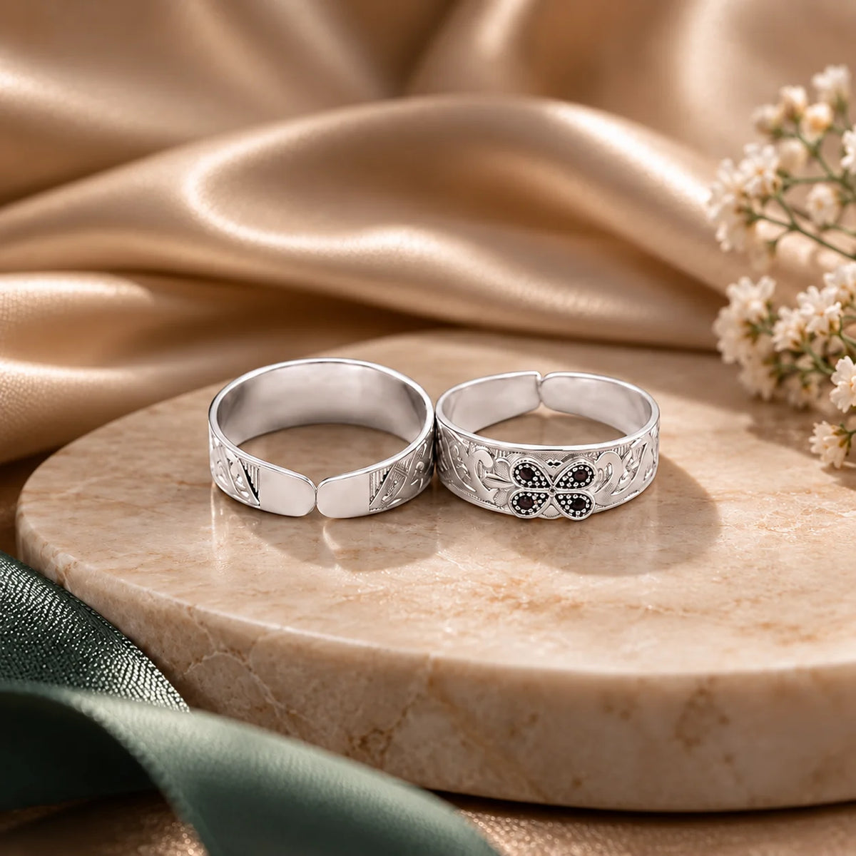 Two silver-toned adjustable rings displayed on a round beige stone slab, with draped champagne satin fabric and small white flowers in the background.