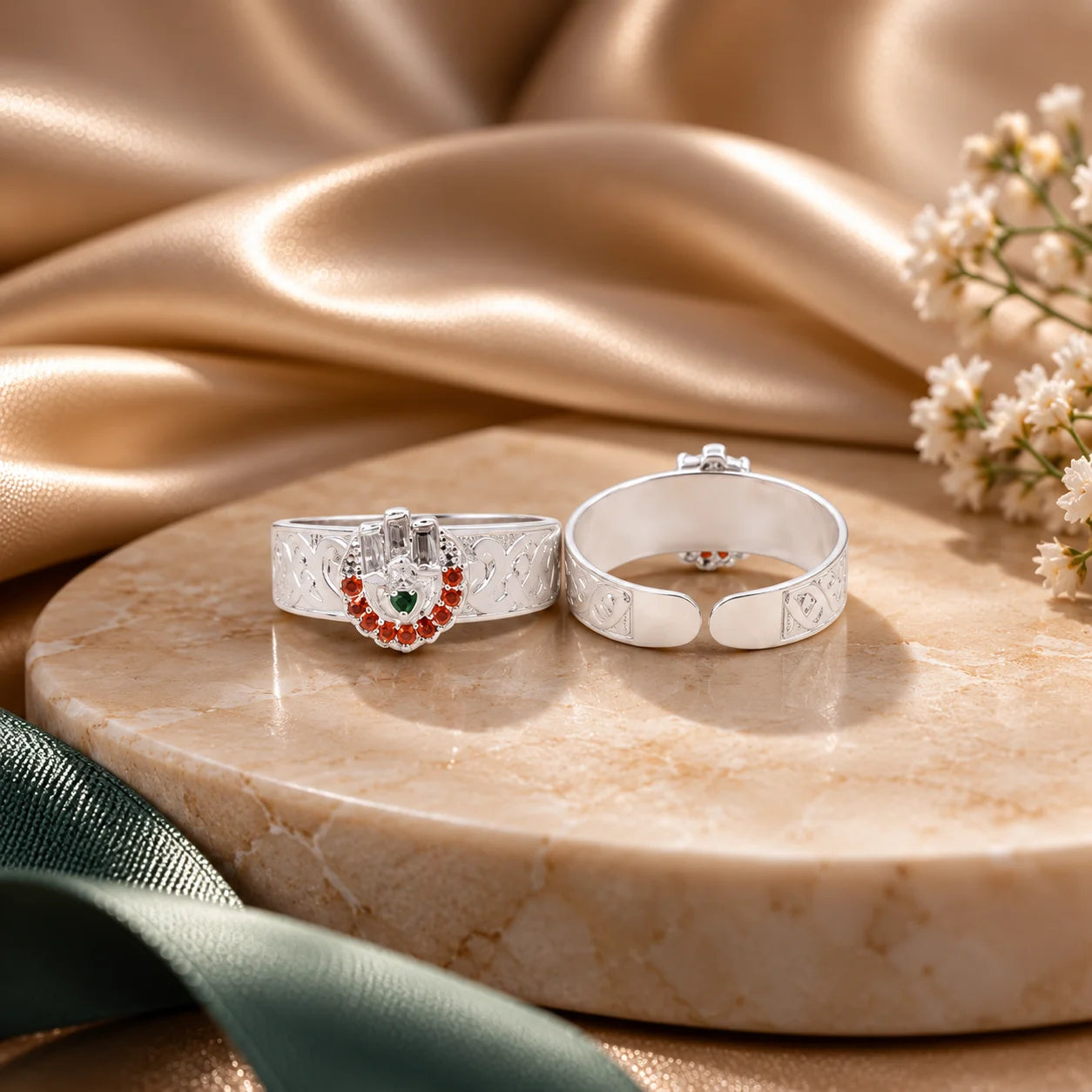 Two silver rings displayed on a round beige stone slab with draped satin fabric and small white flowers. One ring has an engraved floral pattern, and the other features a crescent moon design set with red and green stones.