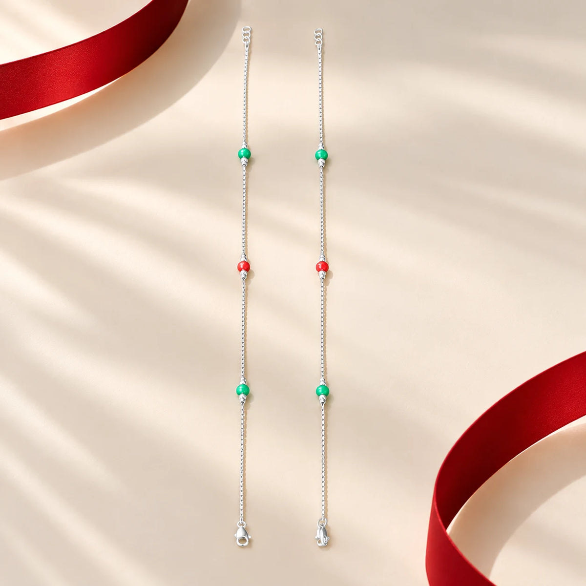 Silver anklet pair with red and green beads on a cream background by Khushbu Jewellers