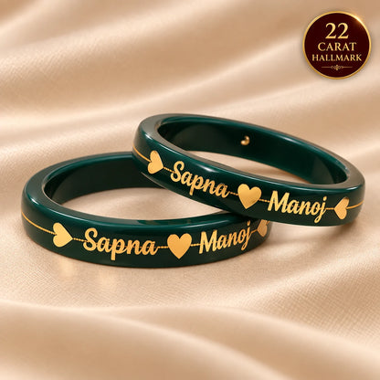 Two dark green bangles with gold engraved names and heart motifs, displayed on a beige satin fabric background. A gold circular badge in the top right corner reads '22 CARAT HALLMARK'.