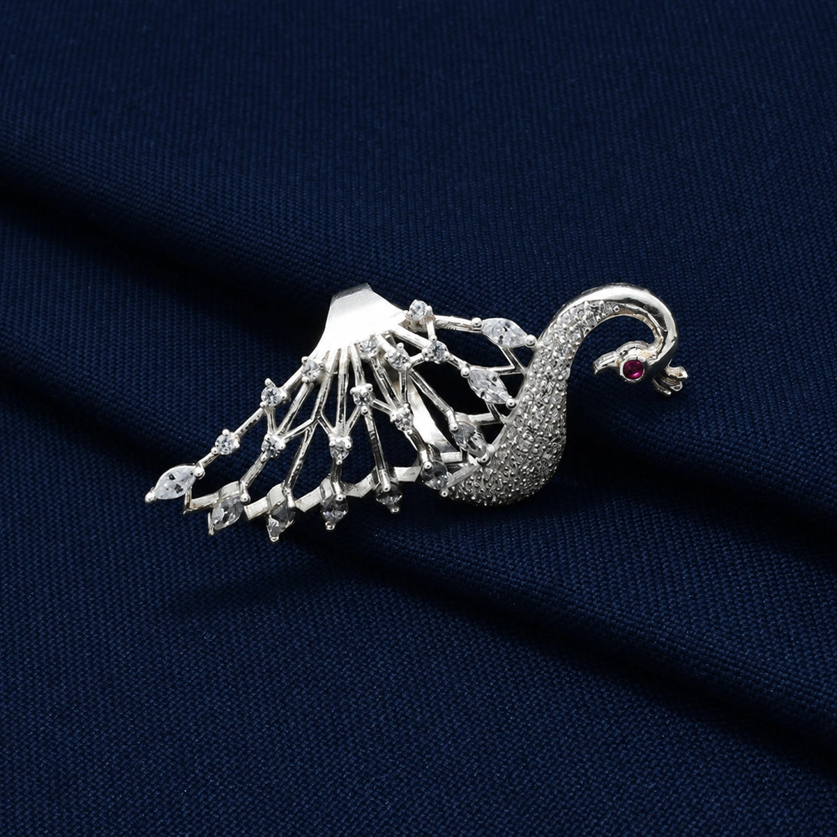 A silver-toned peacock brooch with clear gemstones and a small red eye accent, displayed on a textured dark navy blue fabric background.
