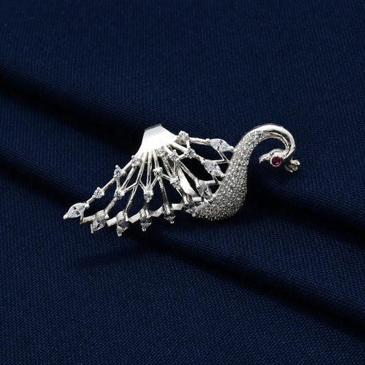 A silver-toned peacock brooch with clear gemstones and a small red eye accent, displayed on a textured dark navy blue fabric background.